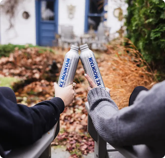 Two people in sweaters clink Ice Mountain water bottles while sitting outdoors, surrounded by autumn foliage and a blurred house in the background.